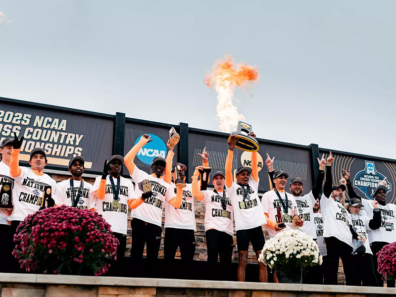 Oklahoma State men’s cross country team celebrates winning the 2025 NCAA national championship, holding trophies and medals on stage with a flame display in the background.