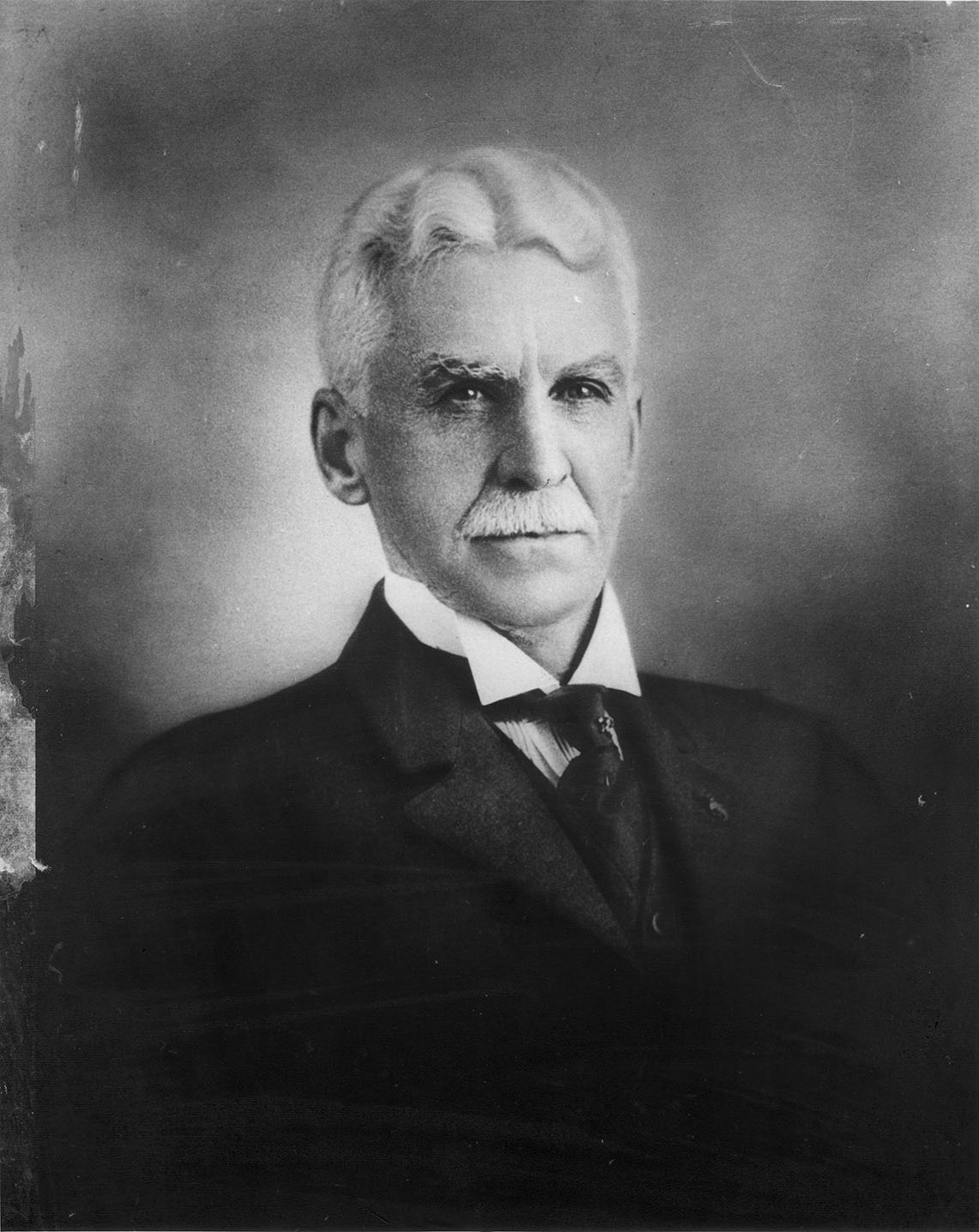 A black and white photograph of a man with short, light-colored hair, dressed in a dark suit, tie, and collared shirt, set against a plain, slightly blurred background.